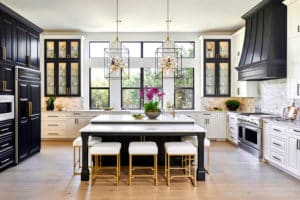 A kitchen with black and white cabinets, white marble countertops, a large island with white cushioned stools with a vase of purple flowers, pendant lighting over the island, and wood floors.