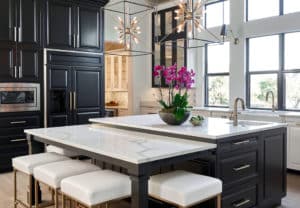 A kitchen with black cabinets, white marble counters, a large island with white cushioned stools with a vase of purple flowers.