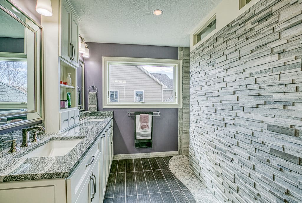 Bathroom with natural stone tile floors in black woodgrain look. Double vanity with stone wall on opposite side. Purple wall at the far end.