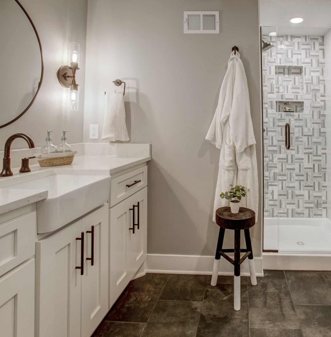 Remodeled bathroom with dual white sinks, with round mirror centered, and gold trim. Also, a glass walk-in shower with a robe hanging